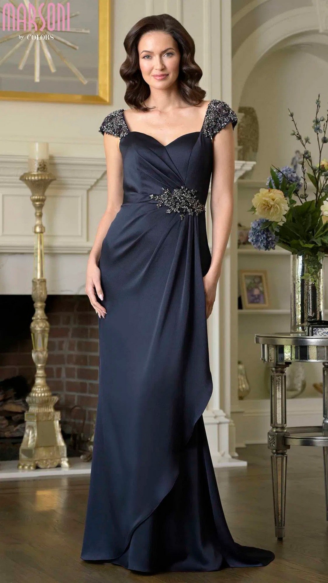 A woman in a navy Marsoni MV1377 long formal dress with beaded cap sleeves and an empire waist stands in a living room near a fireplace and floral arrangements.